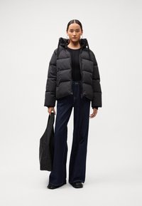 Black puffer jacket with a hood, quilted design, worn over a fitted black top. Paired with high-waisted dark denim jeans and a black tote.