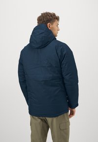 Marineblaue isolierte Jacke mit Kapuze, ausgestattet mit einer glatten Textur und einem leicht verlängerten hinteren Saum. Getragen über olivgrünen Hosen.