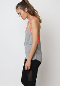 Top gris sin mangas con tirantes naranjas, corte relajado, dobladillo redondeado y una textura suave. Combinado con leggings negros que presentan recortes de malla.