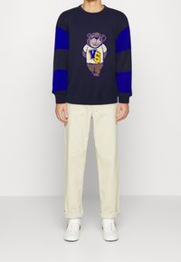 Pull en tricot bleu marine avec des rayures bleues, arborant un motif d'ours portant une chemise blanche avec l'inscription « VS ». Porté avec un pantalon crème et des baskets blanches.