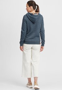 Sweat à capuche bleu avec une doublure texturée beige, associé à un pantalon large blanc et des baskets de couleur claire. Il présente une étiquette en cuir.