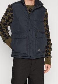 Marinblå puffväst med framknäppningar och två stora fickor, med ståkrage och en slät textur. Buren över en mönstrad tröja.