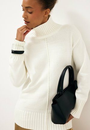 Donna che indossa un maglione bianco a collo alto, tiene in una mano una borsa nera e si tocca il mento con l'altra mano.