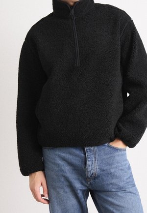 Pull en polaire noir avec une fermeture éclair quart porté par une personne avec une main dans la poche d’un jean bleu sur un fond uni.