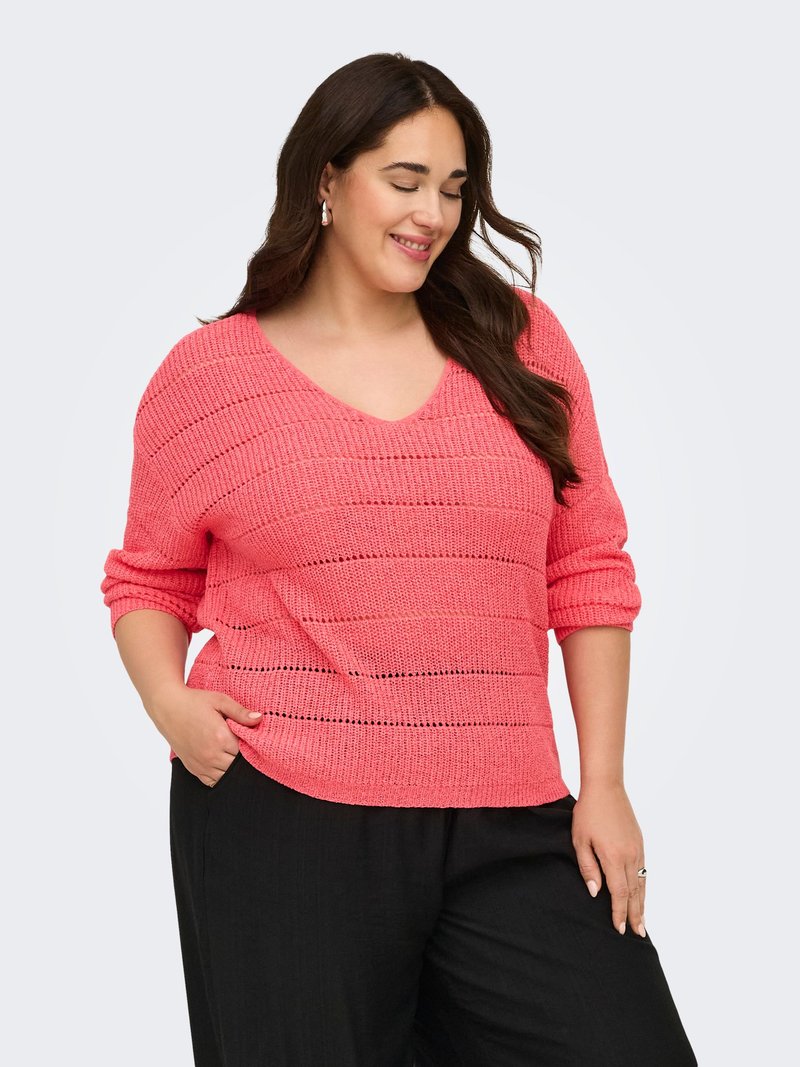 Femme souriante aux longs cheveux foncés portant des boucles d'oreilles en perles, un pull en maille rose ample avec les manches retroussées et un pantalon noir, posant avec une main dans la poche.