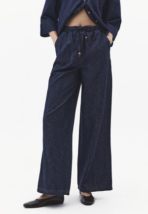 Person iført mørkeblå vidde bukser med elastisk talje og snor, parret med sorte flade sko og en delvist knaplukket skjorte.