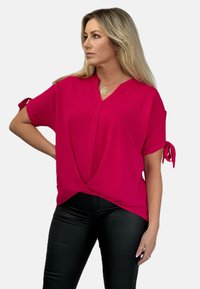 Fuksjowa bluzka z krótkim rękawem, z głębokim dekoltem w kształcie V i wiązaniami na rękawach, w połączeniu z czarnymi dopasowanymi spodniami. Teksturowany, lekki materiał.
