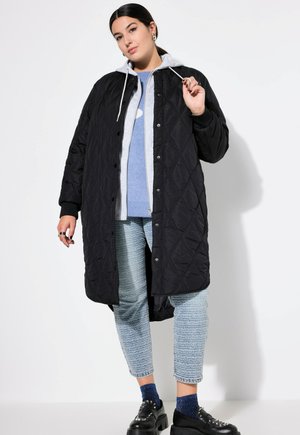 Zwarte gewatteerde lange jas met knoopsluiting en een grijze capuchon, gedragen over een lichtblauwe trui en gestreepte blauwe jeans. Zwarte platform schoenen.