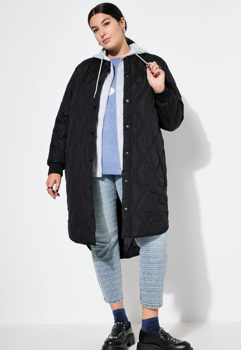 Cappotto lungo trapuntato nero con chiusura a bottoni e cappuccio grigio, indossato sopra un maglione blu chiaro e jeans blu a righe. Scarpe con piattaforma nere.