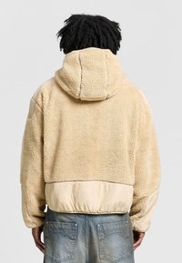 Beige fleecejacka med huva, med en strukturerad yta och släta tygdetaljer vid midjan och ärmarna. Avslappnad design, inga synliga logotyper.