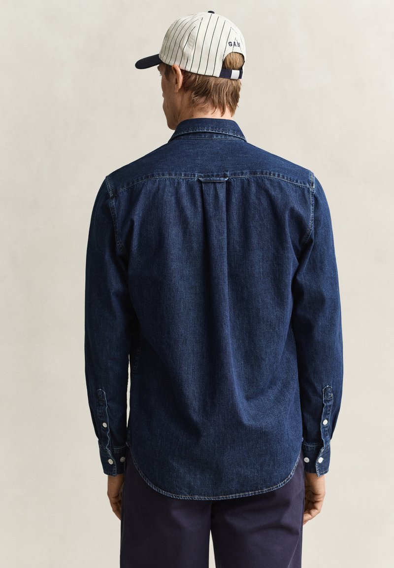 Mann, der von hinten zu sehen ist, trägt ein dunkelblaues Jeanshemd, marineblaue Hose und eine weiße Baseballkappe mit dünnen Streifen und Text auf der Rückseite.