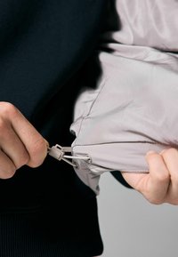 Pilkas zipuotas švarkas su lygiu paviršiumi, turintis reguliuojamą raištelį ir juodą tekstūruotą audinį po juo.