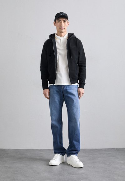 Haut à capuche noir zippé sur un polo blanc, jeans bleus et baskets blanches ; le modèle se tient droit contre un fond gris. Design simple, coupe décontractée.
