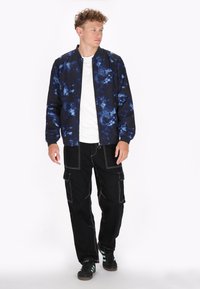 Bomberjack, uitgevoerd in een blauw en zwart tie-dyepatroon, met ribgebreide manchetten en kraag. Gecombineerd met zwarte cargopants en zwarte sneakers met groene accenten.