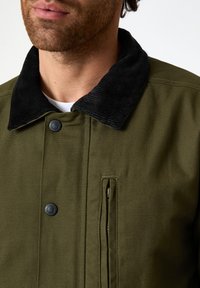 Giacca verde oliva con colletto in velluto a coste nere, chiusura con bottoni a pressione e tasca laterale con zip. Tessuto morbido con design minimalista.