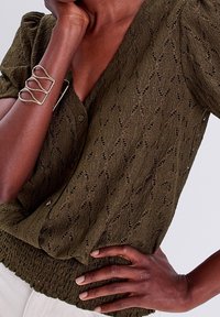 Blouse en maille vert olive avec un col en V, des manches courtes bouffantes et un ourlet côtelé. Présente un motif texturé et une fermeture à boutons.