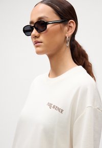 Mujer con el cabello castaño recogido, usando gafas de sol ovaladas negras, aretes de aro plateados y una camisa blanca con el texto "Park Avenue".