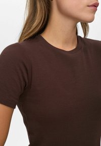 Moteris vilkinti prigludusią trumpomis rankovėmis šokoladinio rudo tono dryžuotą marškinėlį, matyti kaklo šonas, žandikaulis ir lūpos prieš baltą foną.