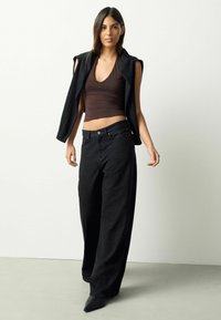 Blugi negri cu talie înaltă și croială largă, asortați cu un top maro mulat și un sacou negru fără mâneci, cu detalii decupate pe umeri.