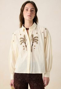 Cream-colored button-up shirt with palm tree and floral embroidery, long sleeves, and a light textured fabric. Paired with patterned dark trousers.