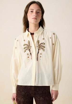 Cream-colored button-up shirt with palm tree and floral embroidery, long sleeves, and a light textured fabric. Paired with patterned dark trousers.