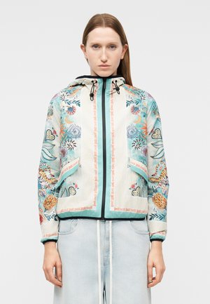 Vrouw draagt een jas met capuchon en bloemenpatroon, met voorzakken en lichtblauwe broek tegen een effen achtergrond.