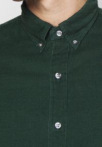 Tmavě zelená kordurová košile s límečkem na knoflíky a stříbrnými knoflíky, s texturovaným žebrovaným vzorem. Detailní záběr.