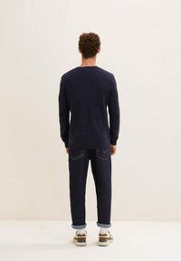 Man med lockigt hår som vänder bort ansiktet, iförd en marinblå prickig tröja, mörka uppvikt jeans, vita strumpor och brunvita sneakers.