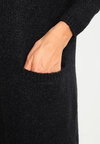 Czarny sweter dzianinowy z prążkowanym mankietem i kieszenią z przodu, prezentujący miękką teksturę i dopasowany krój.