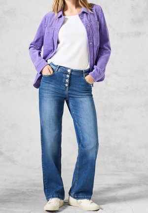Chemise en velours côtelé violet, haut blanc à col rond, jean bleu à jambes larges avec fermeture boutonnée, porté avec des baskets blanches, sur un fond gris clair.