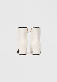 Bottes blanches à la cheville en matériau lisse avec un talon carré, dotées d'une doublure intérieure noire et d'une fermeture éclair dorée à l'arrière.