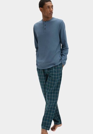 Mann steht barfuß, trägt ein blaues Henley-Shirt mit langen Ärmeln und türkis karierte Hose, blickt nach links mit den Händen in den Taschen.