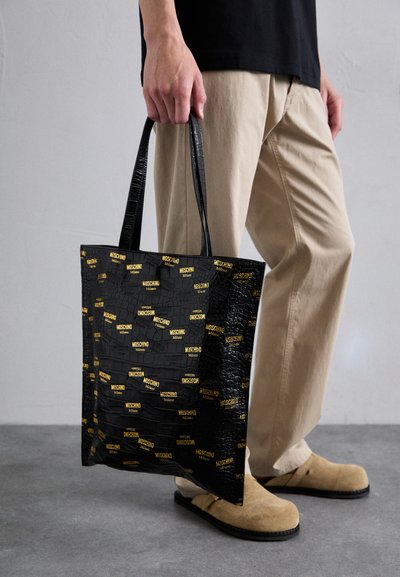 Geantă de umăr neagră cu textură de crocodil, având branding galben "Moschino". Mânerile sunt drepte și realizate din același material lucios.