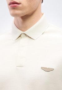 Κοντινό πλάνο ενός ατόμου που φορά λευκό πόλο πουκάμισο με γιακά, με ένα σήμα λογότυπο Aston Martin στο στήθος.