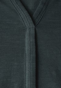 Tissu côtelé vert foncé avec un design à col en V ; présente des coutures subtiles le long du col et de la patte de boutonnage pour le détail. Texture douce.