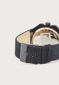 Reloj de pulsera textil negro con correa tejida, que presenta una caja cuadrada negra, detalles en plata y un lado inferior de cuero. El cierre muestra el logotipo de la marca.