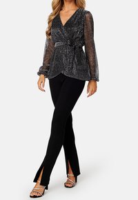 Haut cache-cœur noir avec manches longues transparentes et texturées, noué à la taille, associé à un pantalon noir ajusté avec des ourlets fendus et des talons hauts.