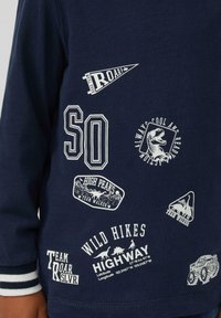 Marineblauw long-sleeve shirt met witte grafische prints. Voorzien van verschillende patches met een buitenthema, waaronder bergen en dinosaurussen.