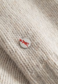 Beige gebreide stof met een een gestructureerd patroon, voorzien van een ronde metalen label met een uitgesneden rode hondensilhouet bevestigd met rode draad.