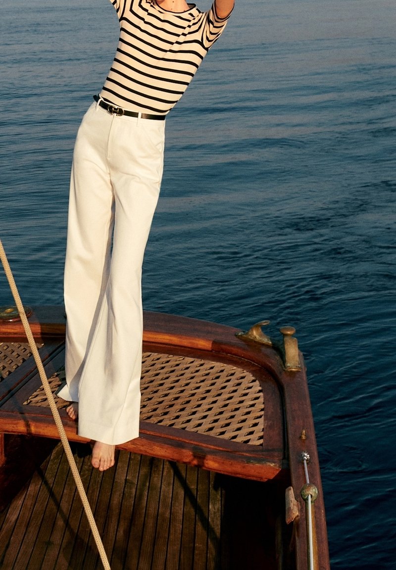 Personne en chemise rayée et pantalon blanc debout pieds nus sur le pont en bois d’un bateau près d’une eau calme.