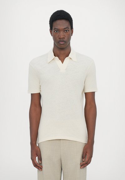Polo beige clair avec une finition texturée, présentant un col et des manches courtes. Associé à un pantalon de couleur claire.