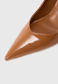 Zapato de mujer de cuero marrón brillante con punta puntiaguda y costuras visibles a lo largo del borde superior sobre un fondo claro y sencillo.