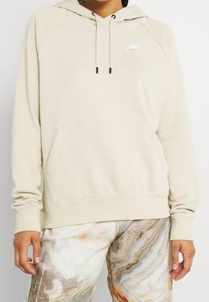 Personne portant un sweat à capuche Nike beige avec cordons et un pantalon à motif marbré dans des tons neutres et orange.