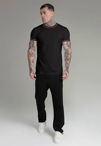 Fekete rövid ujjú póló finoman kontrasztos gallérral, fekete nadrággal és fehér tornacipővel párosítva, mindez testhez álló stílusban.