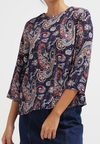 Mörkblå, lätt blus med paisleymönster i röd, vit och grön. Har en rund hals och trekvartslånga ärmar.