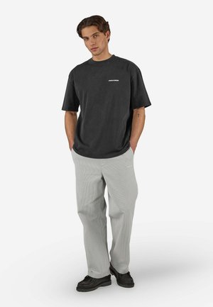 Ung mand iført en sort Pegador T-shirt, lysegrå bukser og sorte sko, stående med hænderne i lommerne på en hvid baggrund.