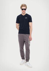 Námornícky modré tričko s výstrihom do V a logom; šedé tepláky s bielou grafikou; biele tenisky; model tiež nosí slnečné okuliare.