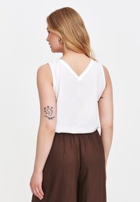 Biała bezrękawnikowa bluzka z dekoltem w serek i miękkiego materiału, w połączeniu z brązowymi spodniami na wysokim stanie. Ma tatuaż na lewym ramieniu.