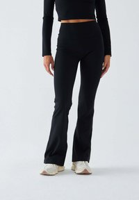 Czarne legginsy z rozszerzanymi nogawkami z elastycznego materiału, z wysokim stanem, o gładkiej teksturze, zestawione z białymi i beżowymi butami sportowymi.