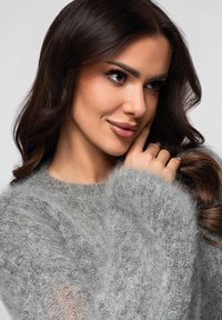 Jeune femme aux longs cheveux foncés portant un pull gris duveteux, posant sa main sur sa joue et regardant légèrement sur le côté.